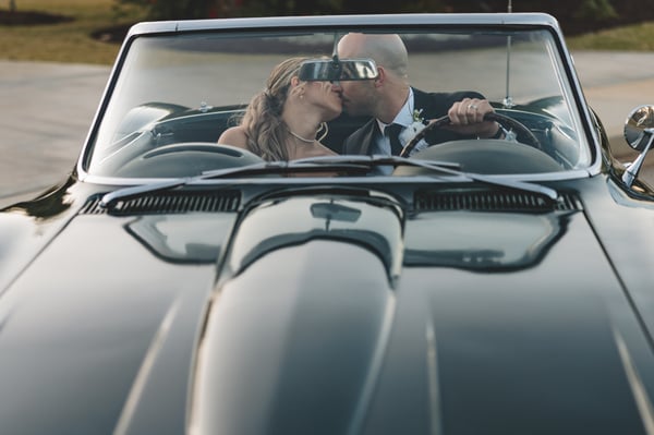 Front-on view looking over the Corvette hood as the bride and groom kiss inside, framed by the windshield and soft dusk light