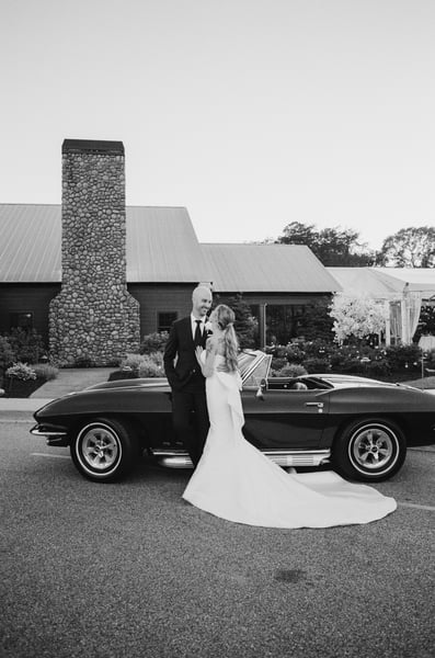 Bride and groom standing beside the vintage Corvette at dusk, stone chimney venue building behind them