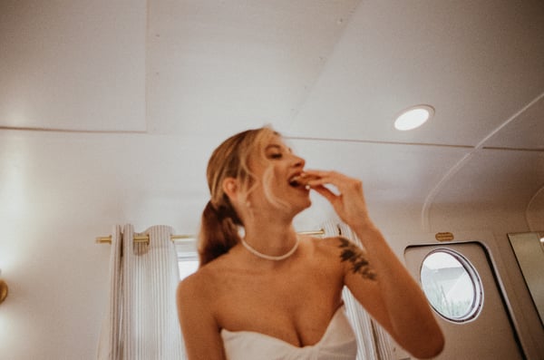 Bride in a strapless gown laughing while taking a bite of food, soft window light through a porthole behind her