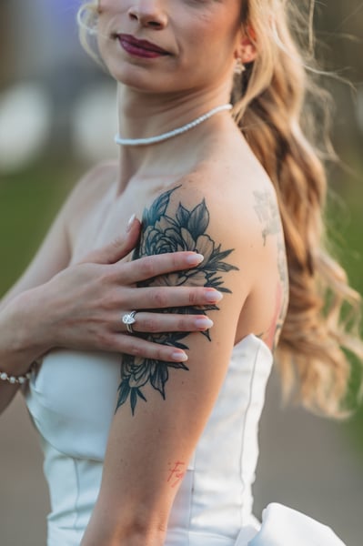 Close-up of the bride's floral arm tattoo, her hand resting on her shoulder, engagement ring visible, pearl choker below