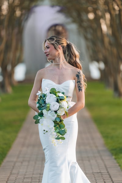 Bride walking along a tree-lined brick path carrying a cascading white bouquet, looking to the side