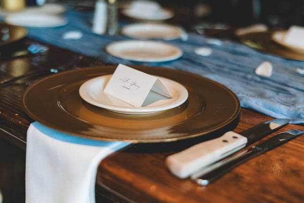 Wedding place setting with a gold charger plate, white dinner plate, and a calligraphed escort card, with a blue linen runner on a wooden table