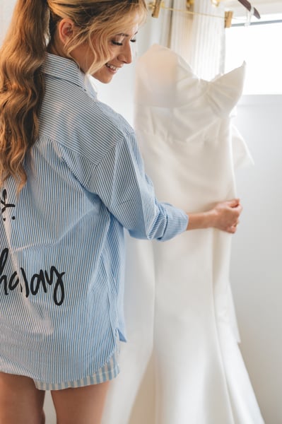 Bride in a blue-striped getting-ready shirt smiling as she touches her hanging wedding gown by a sunlit window