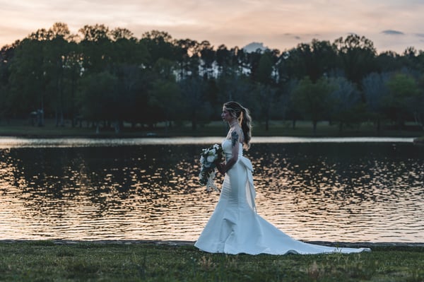 Wide landscape shot of bride walking along the lake at dusk, bouquet at her side, soft pink sky and still water behind her