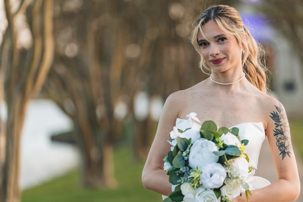 Close-up three-quarter portrait of bride with bouquet at chest height, golden hour trees behind her, looking directly at camera
