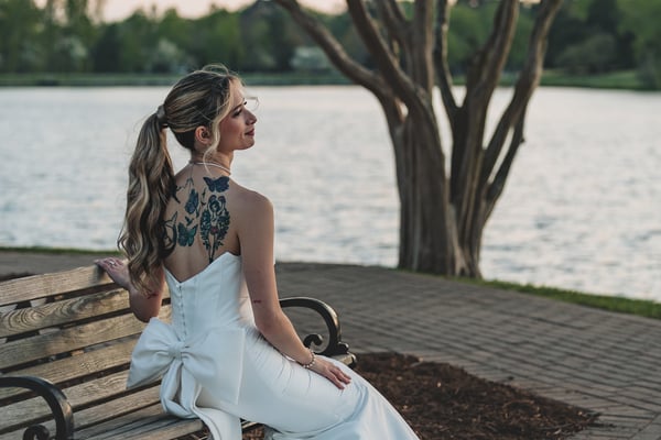 Bride seated on a lakeside bench, back to camera, colorful back tattoos fully displayed, gown and bow draped over the seat, dusk light
