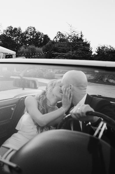 Bride and groom kissing inside the Corvette, captured through the windshield with string lights glowing behind them