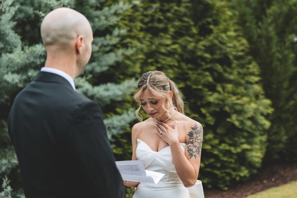 Bride overcome with emotion, hand pressed to her chest, tears streaming as she listens to the groom read his vows from a card in the foreground