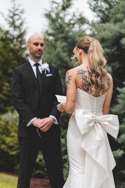 Bride facing the camera, reading her vows from a card with a steady gaze, while the groom listens in soft focus behind her, evergreen trees surrounding them