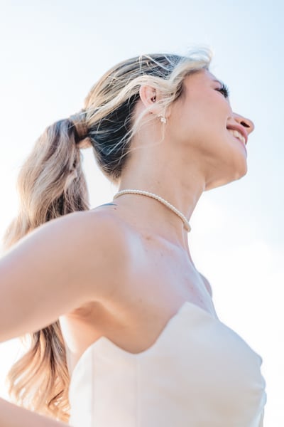 Low-angle close-up portrait of bride's neck and shoulder, pearl choker necklace and earring visible, bright sky behind