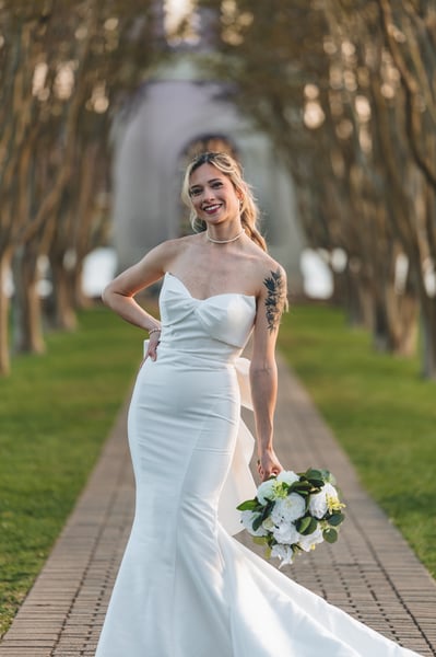 Bride facing camera on the tree-lined brick alley, hand on hip, bouquet at her side, confident smile, afternoon light