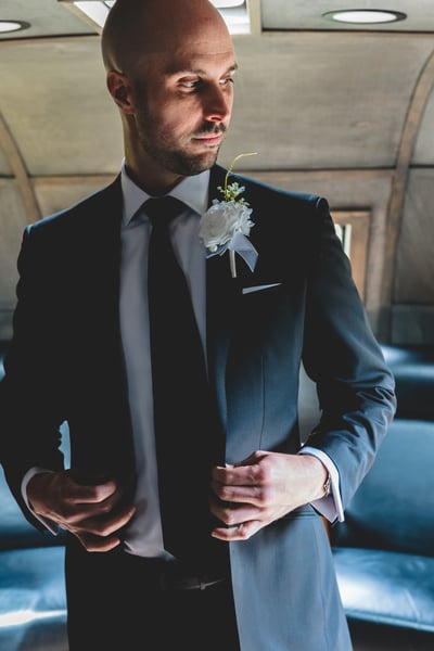 Groom in a charcoal suit with white boutonniere standing in a moody interior setting, adjusting his jacket and gazing to the side