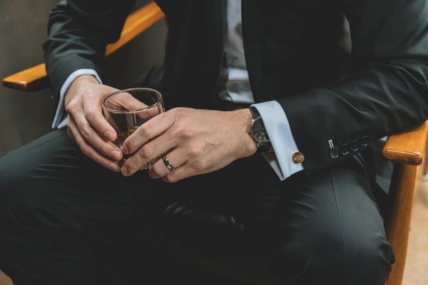 Groom seated holding a crystal glass of whiskey, his watch and gold cufflinks on display against his dark suit