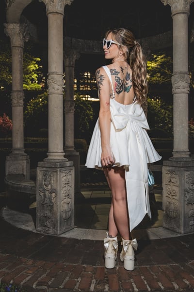 Bride in short white dress from behind inside the lit night gazebo, bow back and colorful tattoos visible, blue bag dangling from her hand
