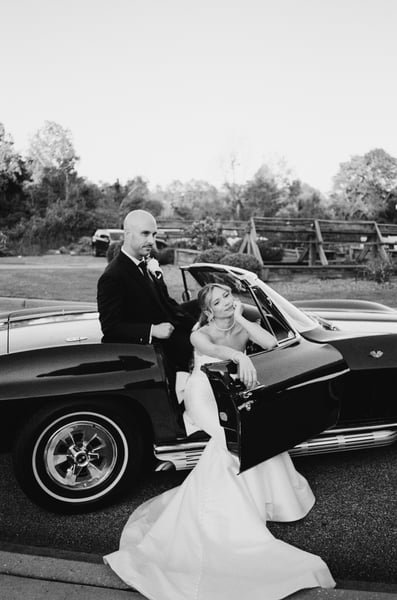 Groom standing behind the Corvette while bride leans out the open car door and looks up at him