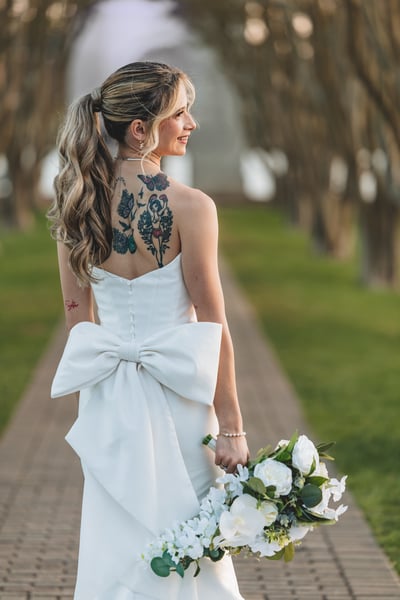 Bride on the tree-lined path, back showing the gown bow and colorful back tattoos, smiling over her shoulder holding bouquet