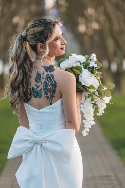 Bride standing centered on a symmetrical brick tree-lined path with a fountain or structure in the distance, full-length portrait with bouquet