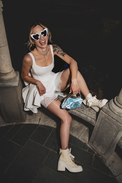 Bride in short white dress laughing freely in the night gazebo, full-length, heart sunglasses, lace garter peeking below the hem, blue bag and Converse on display