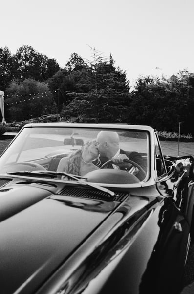 Bride and groom kissing in the Corvette, photographed through the windshield with string lights glowing behind them