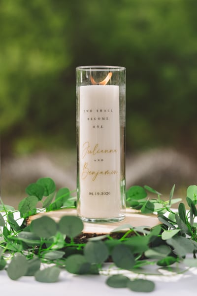 Ceremony unity candle etched with Julianna and Benjamin's names and the date 04.19.2026 and the phrase Two Shall Become One, resting on a wood slice surrounded by greenery