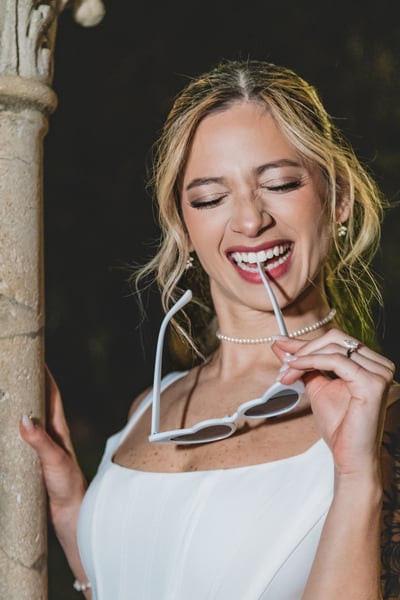 Close-up portrait at night of bride playfully biting the arm of her heart-shaped sunglasses, pearl choker and ring visible, laughing