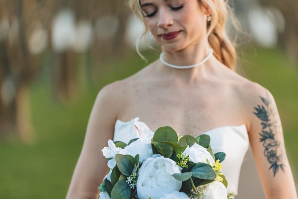 Close-up of bride's face and bouquet, eyes gently closed, white peonies and greenery filling the frame below her pearl choker