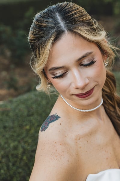 Close-up portrait of bride with eyes downcast, pearl choker necklace, soft blush makeup, boxwood hedge out of focus behind her