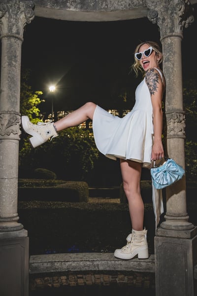 Bride in short white dress kicking one leg up playfully in the night gazebo, heart sunglasses, bejeweled Converse, blue bow bag