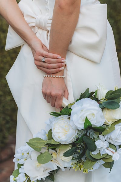 Close-up of bride's hands resting on the back bow of her gown, engagement ring and pearl bracelet visible, bouquet of white peonies below