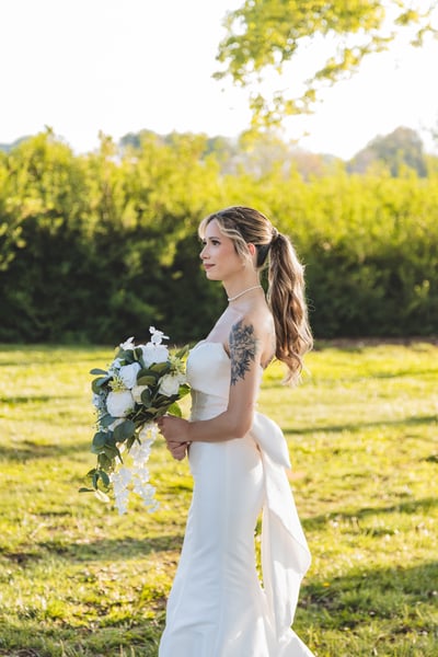 Three-quarter portrait of bride in open park field, golden-hour backlight, bouquet held out, looking over her shoulder with a soft smile