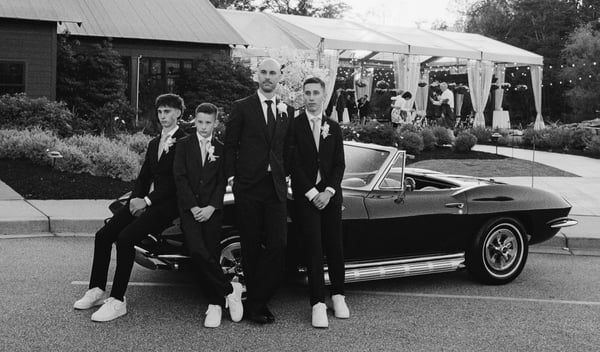 Groom standing with three junior groomsmen in suits and boutonnieres leaning on the Corvette, reception tent glowing behind them