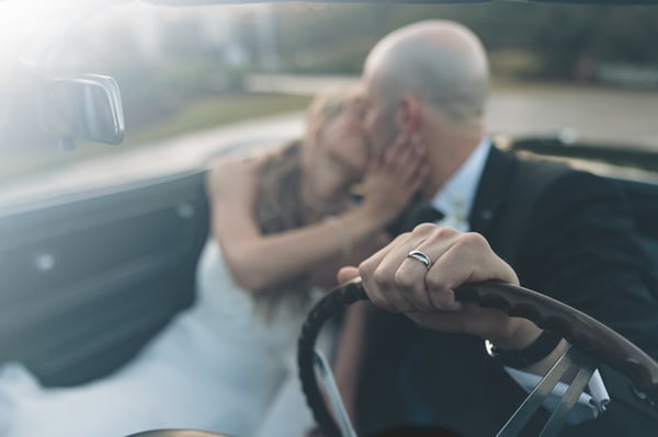 Close-up of the groom's wedding band on the Corvette steering wheel in sharp focus, with the couple sharing a soft kiss in the blurred background