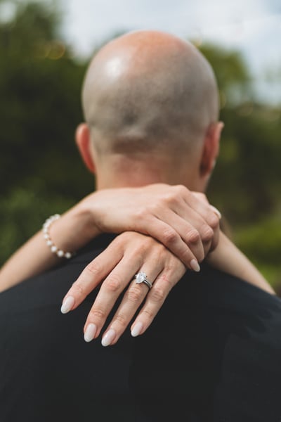 Bride draping her arms around the groom's neck from behind with her ring hand visible, shot from behind showing the groom's head and the outdoor garden background