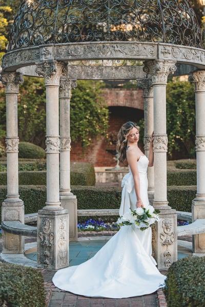 Bride in mermaid gown in side profile inside the iron-dome garden gazebo, gown train sweeping behind her, bouquet held low