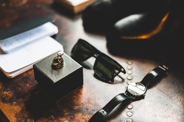Groom's getting-ready flat lay showing gold cufflinks in a box, sunglasses, a watch, and dress shoes arranged on a wooden surface