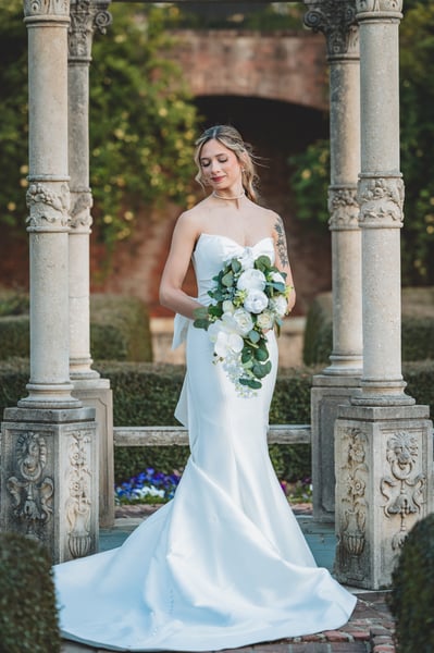 Bride framed between two stone columns inside the formal garden gazebo, blue pansies and boxwood hedges behind her, bouquet held low
