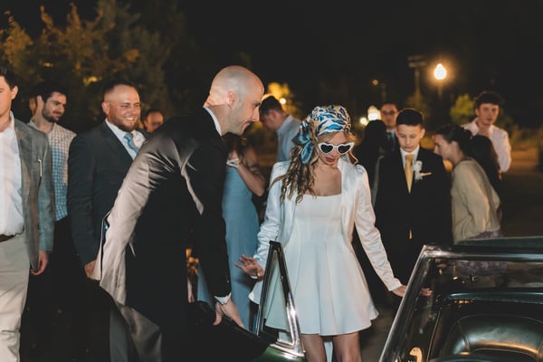 Groom helping the bride into the passenger seat of the Corvette at the sparkler send-off, guests lining the path, bride in heart sunglasses and blue silk headscarf
