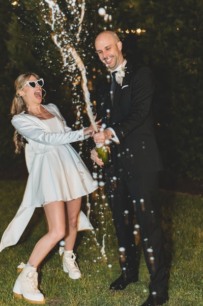 Bride and groom walking through a cheering crowd of guests outside at night, she wearing heart sunglasses and her white mini reception outfit, guests waving sparklers and phones