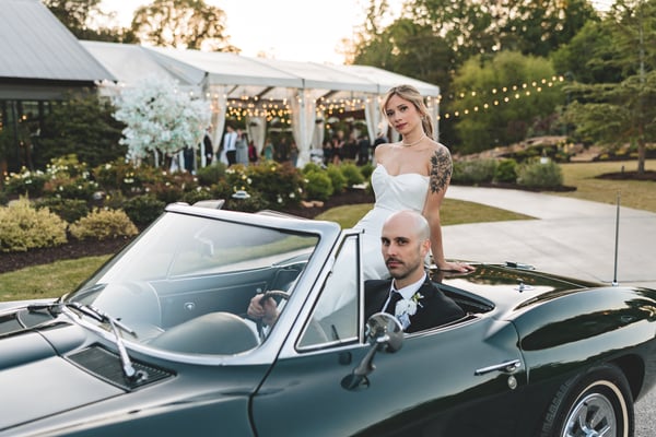 Groom at the wheel of the dark green vintage Corvette, bride standing through the open top beside him, both posed coolly at golden hour with the reception tent and gardens behind