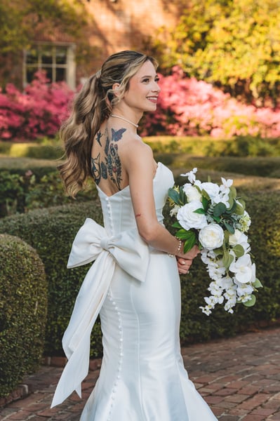 Bride walking away on a brick garden path, colorful back tattoos visible above the gown bow, bouquet at her side, azaleas and hedges behind