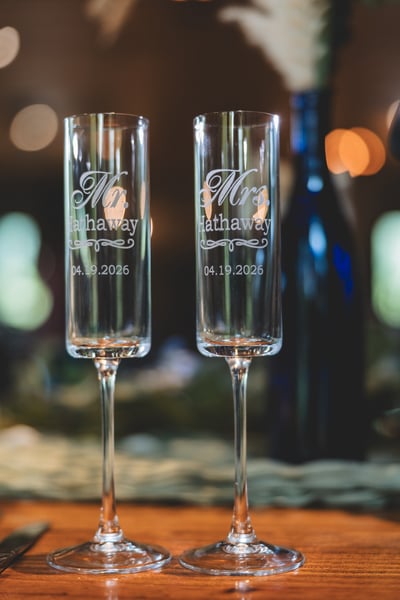 Two engraved crystal champagne flutes inscribed Mr. and Mrs. Hathaway with the date 04.19.2026, standing on a wooden table with bokeh reception lights in the background