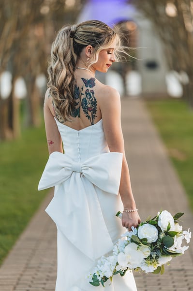 Bride on the tree-lined brick path, back turned, large gown bow and colorful tattoos fully visible, bouquet held loosely