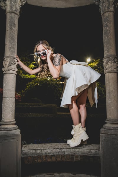 Bride in short white dress leaning between gazebo columns at night, peering forward playfully in heart-shaped sunglasses