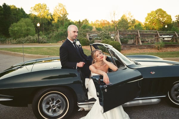 Wide shot of the bride and groom beside the Corvette at dusk, a long wood fence stretching behind them toward the tree line