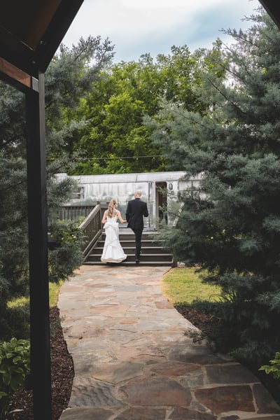 Bride and groom walking away together along a stone garden path toward a vintage airstream trailer, framed by tall cedar trees