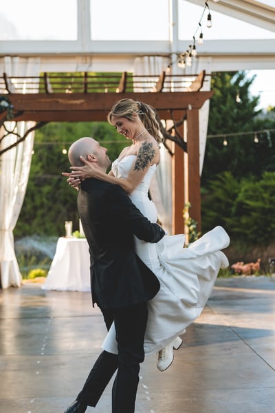 Groom lifting the bride off the ground on the outdoor dance floor under string lights and a pergola, both laughing as they share their first dance