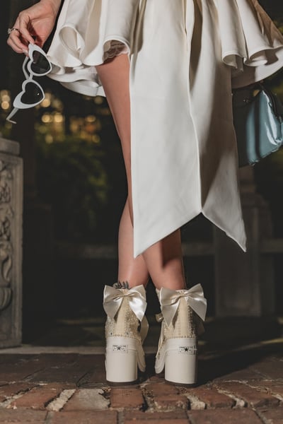 Detail of the backs of both bejeweled platform Converse sneakers with white satin bows, short white dress and blue bag in background