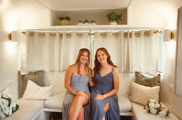 Two bridesmaids in dusty-blue gowns sitting side by side on the airstream bench seat, smiling, with striped curtains and potted greenery behind them