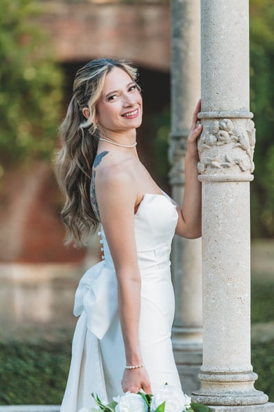 Bride leaning against an ornate stone column in a formal garden, smiling over her shoulder in golden light