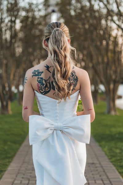 Back of bride on the brick tree-lined path, back tattoos and the large gown bow facing the camera as she looks to the side